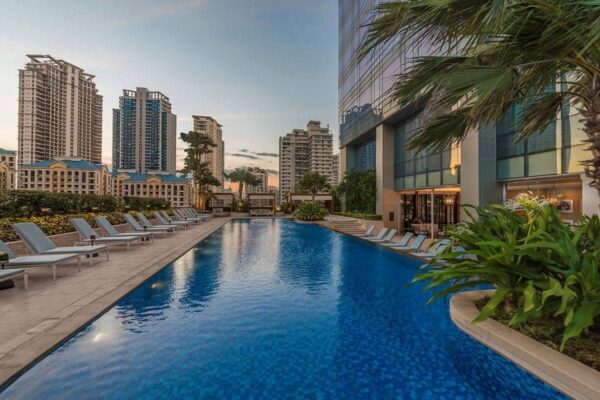 Luxury outdoor swimming pool surrounded by lounge chairs, tropical landscaping, and high-rise condominiums at dusk.